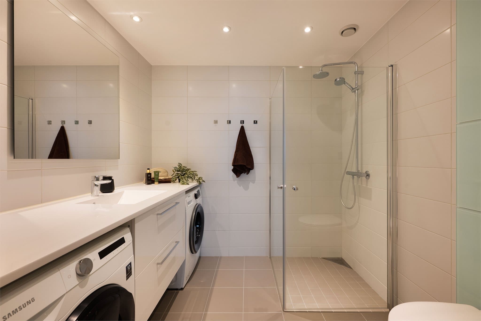 Pent baderom med tidløse fliser med vannbåren gulvvarme for ekstra komfort. Badet har et stilrent servantskap med glatte, folierte fronter og heldekkende servant. Over servant er det speil montert på vegg med lysarmatur.