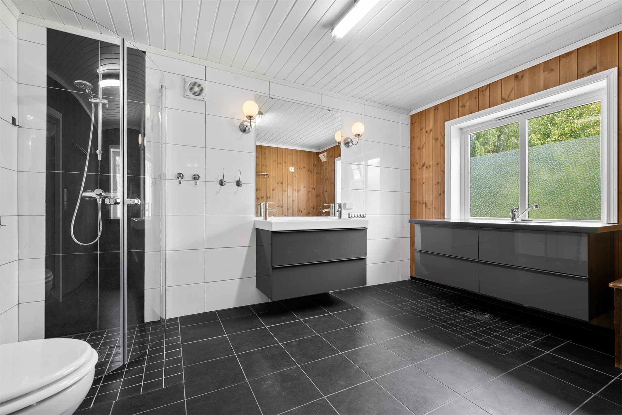 Badet er romslig utformet og fungerer samtidig som et praktisk vaskerom. Her er det god gulvplass, dusjhjørne, vegghengt toalett og moderne baderomsinnredning med servant. Lang innredning med benkeplate, skapplass og integrert stålvask.