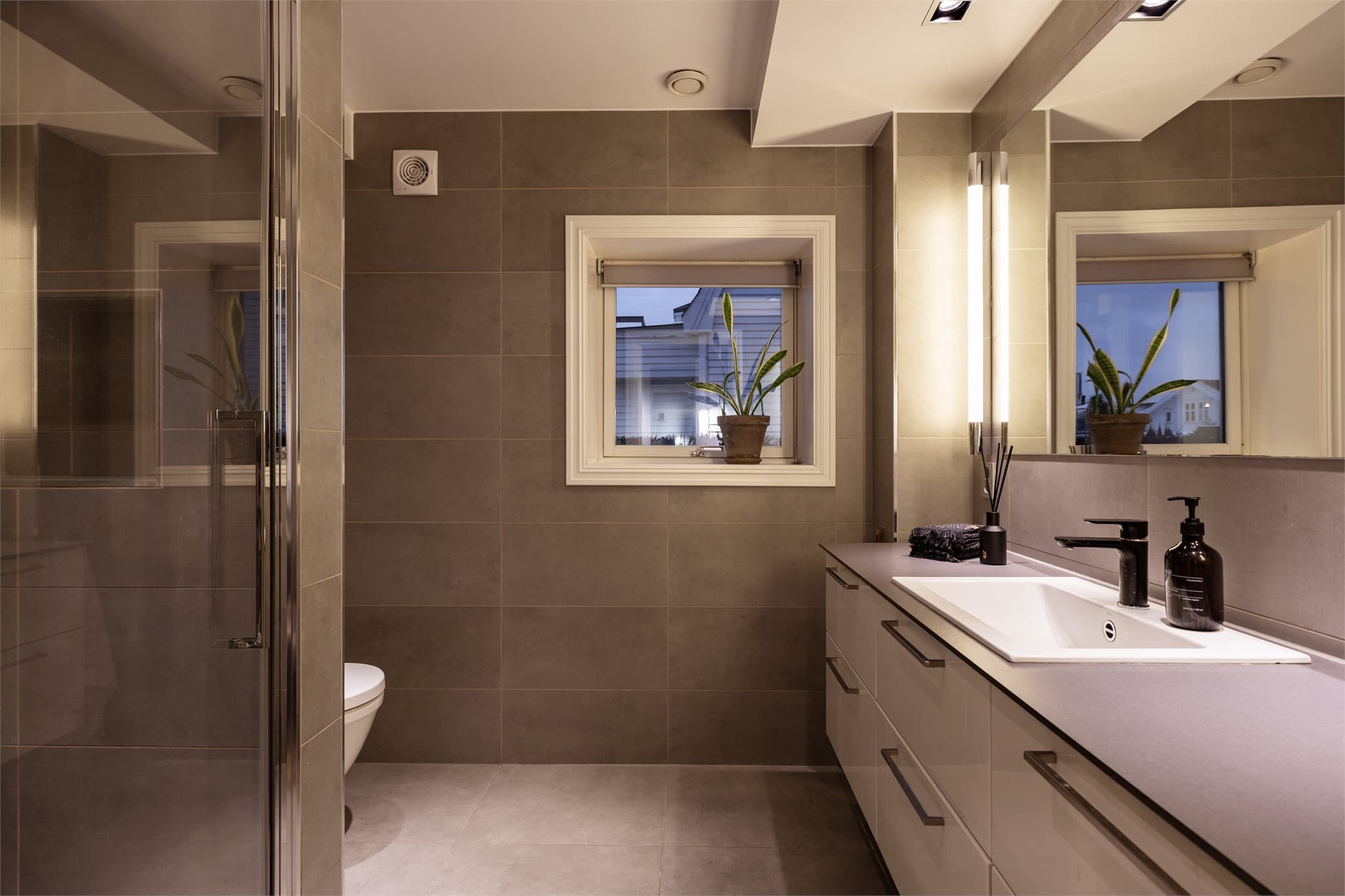 Badet i første etasje fremstår som stilrent og praktisk. Rommet har grå fliser på gulv og vegger, vegghengt toalett, moderne baderomsinnredning med høyglansfronter, servant med speil og belysning, samt en dusjsone.