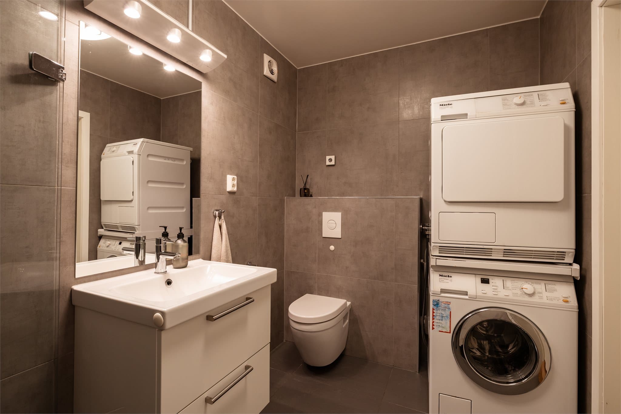 Badet i hybelen er utstyrt med flislagte overflater, moderne baderomsinnredning med glatte fronter, dusj med innfellbare glassdører, servant og vegghengt toalett med innebygd sisterne. Det er også opplegg for vaskemaskin.